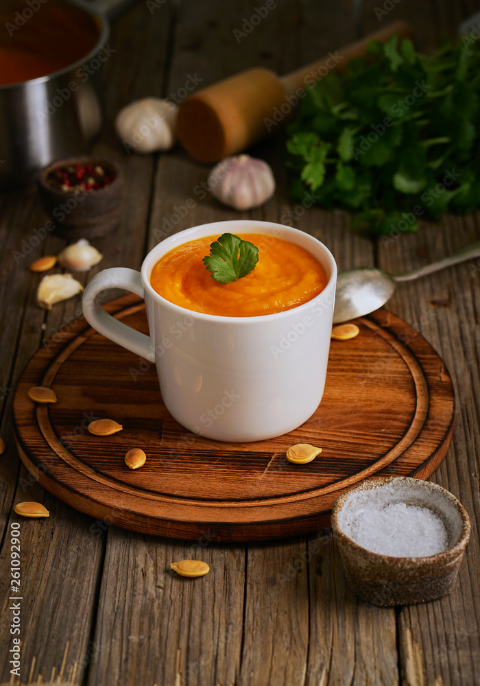 Pupmkin cream soup in cup on brown wooden table, vertical, side view. Dietary vegetarian puree on cutting board with parsley, garlic.