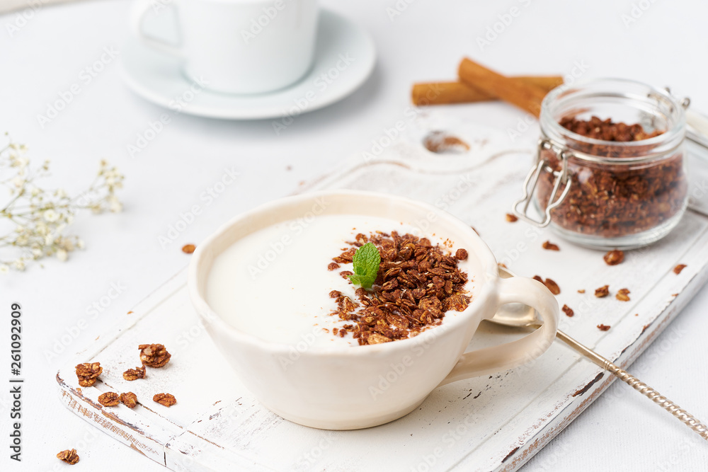 Yogurt with chocolate granola in cup, breakfast with tea on white wooden background, side view. Yogurt with chocolate granola in cup, breakfast with tea on white wooden background, side view.