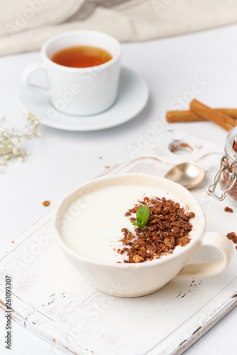 Yogurt with chocolate granola in cup, breakfast with tea on white wooden background, vertical.