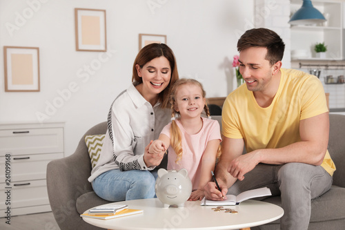 Happy family putting coin into piggy bank at table indoors. Saving money