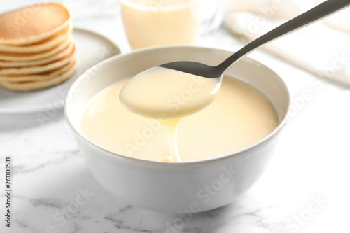 Spoon of pouring condensed milk over bowl on marble table, closeup. Dairy products