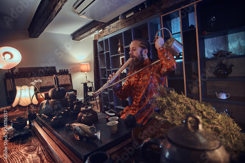 Tea master in kimono making natural tea and playing on a bamboo flute, performs in the dark room with a wooden interior.