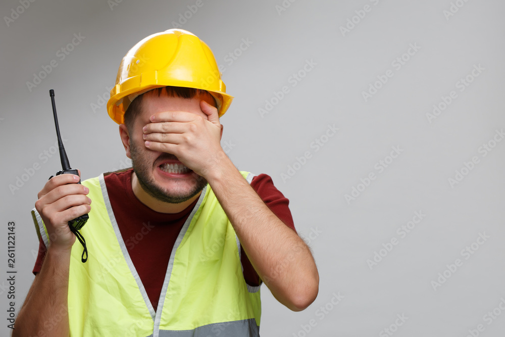 Image of upset builder man in yellow helmet with walkie-talkie on empty gray background. Image of upset builder man in yellow helmet with walkie-talkie on empty gray background.