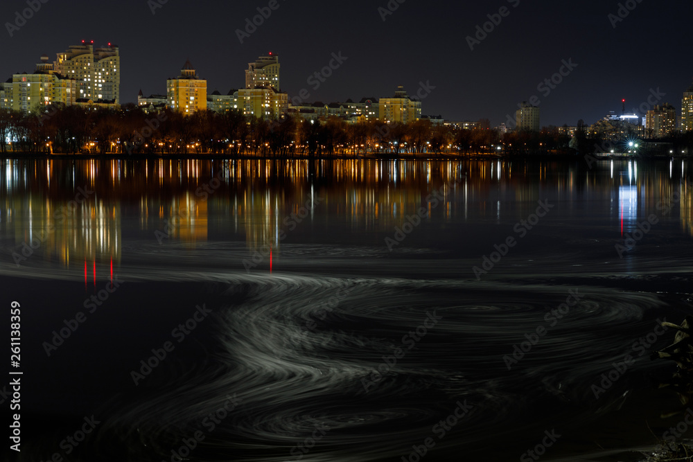 Obraz premium dark cityscape with illuminated buildings, waves on river and night sky