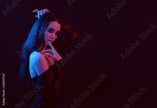 Neon light. Portrait of sexy elegant  beautiful brunette young woman in evening fashionable stylish black dress. Model with trendy evening smoky eyes makeup touching her lips. Dark background.