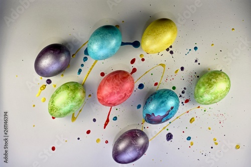 Multi-colored painted eggs on a light surface filled with drops of multi-colored paint. View from above. With shadows.