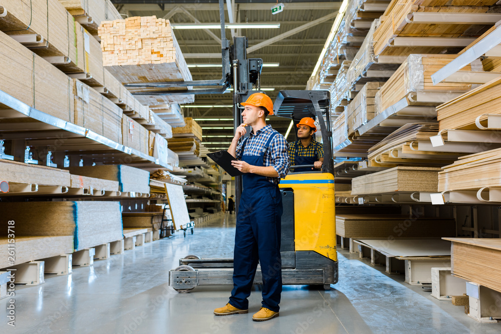 indian warehouse worker sitting in forklift machine near thoughtful colleague with clipboard indian warehouse worker sitting in forklift machine near thoughtful colleague with clipboard