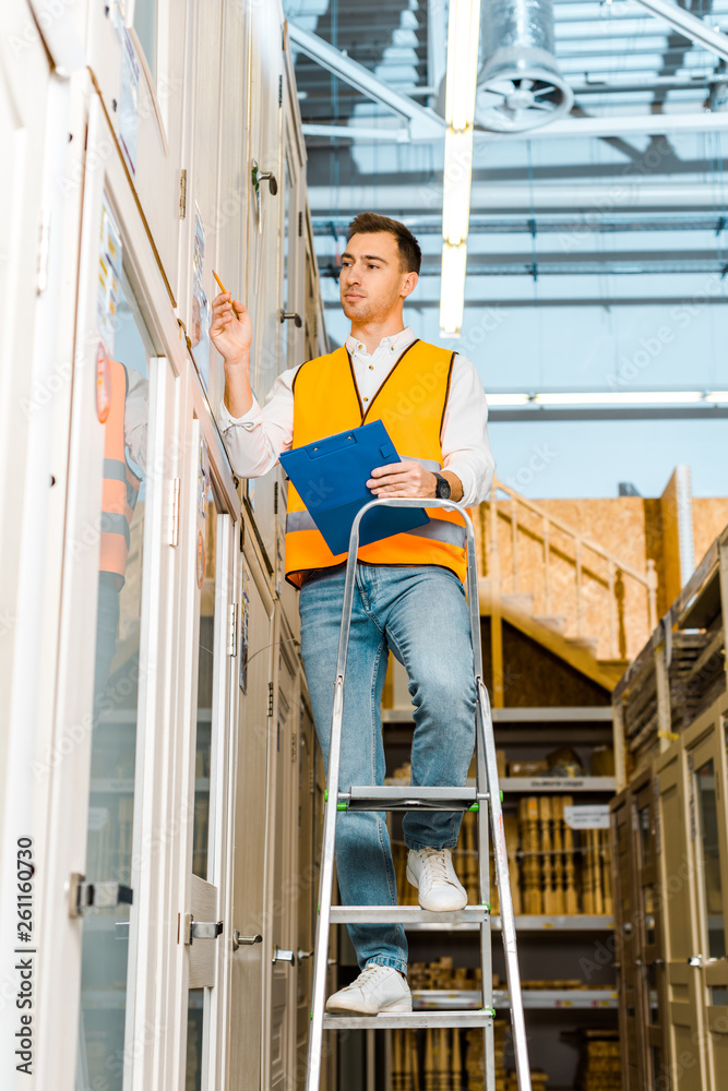 selective focus of concentrated worker in safety vest standing on ladder in doors department