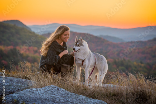 Woman smiles looking her husky dog sitting on an open ridge with the sun rising behind her and mountain ridges.