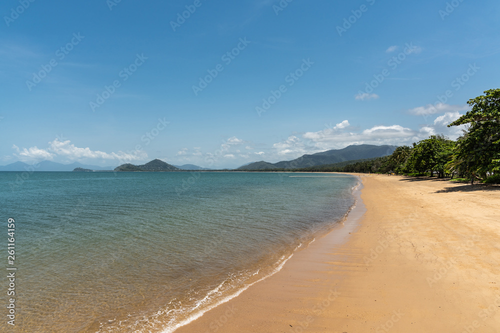Cairns, Australia - February 18, 2019: Warm beige tropical beach of Palm Cove with azure Coral Sea water under blue sky with rainforested mountains on horizon. Cairns, Australia - February 18, 2019: Warm beige tropical beach of Palm Cove with azure Coral Sea water under blue sky with rainforested mountains on horizon.
