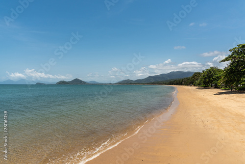 Cairns, Australia - February 18, 2019: Warm beige tropical beach of Palm Cove with azure Coral Sea water under blue sky with rainforested mountains on horizon.