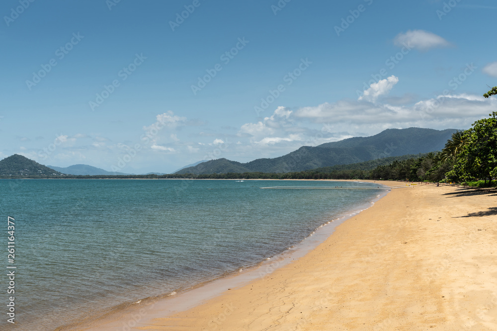 Cairns, Australia - February 18, 2019: Warm beige tropical beach of Palm Cove with azure Coral Sea water under blue sky with rainforested mountains on horizon.