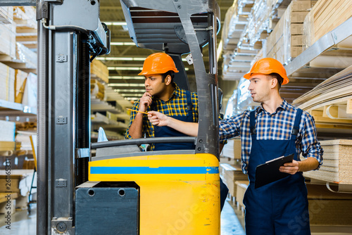 indian warehouse worker sitting in forklift machine near colleague pointing with pencil