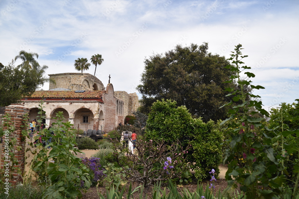 Obraz premium San Juan Capistrano, CA., U.S.A. Ap. 21, 2018. Mission courtyard, fired red brick pillared portico, fountains & water lilies, packed dirt, flat red brick-set paths, beautiful palms, trees & floral 