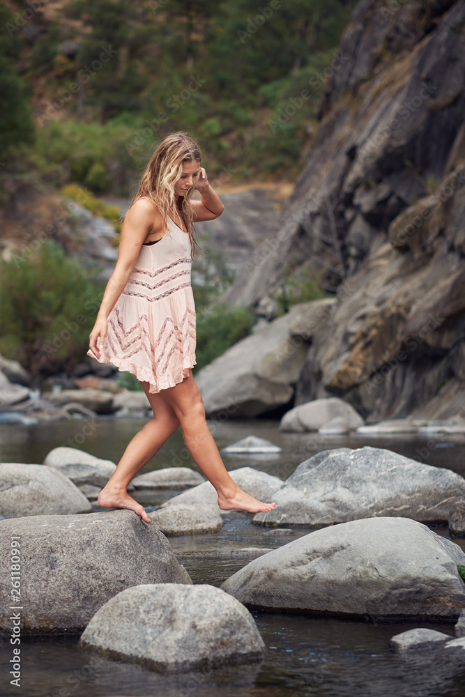 A young, barefoot woman in a summer dress walking across a mountain creek  on rocks. Stock-Foto | Adobe Stock