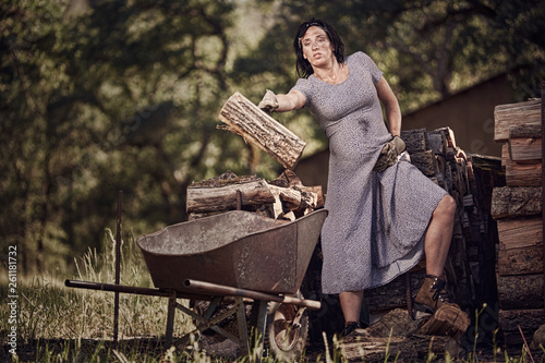 A pergnant woman in a dress throwing wood into a wheelbarrow on a ranch in Northern California.