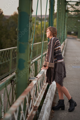 Fashionable boho teenage girl leaning against the rail of a bridge dressed in boots, a dress and sweater.