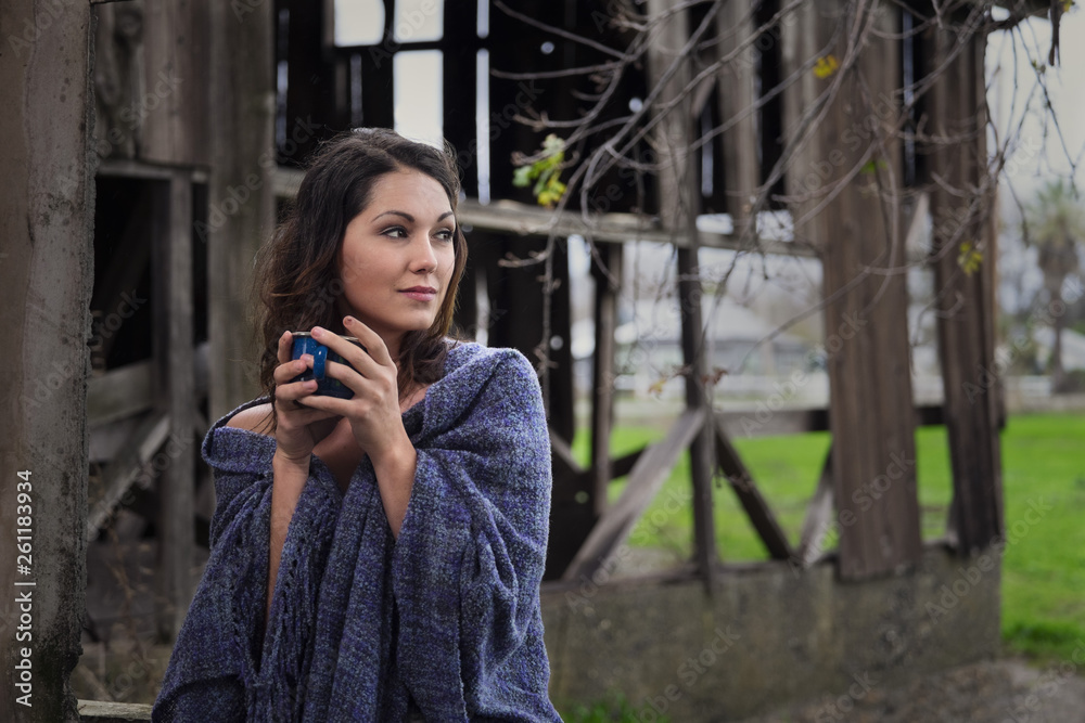 Fototapeta premium A farm girl wrapped in a blue shawl drinking a cup of coffee next to an old barn on a small rural farm.