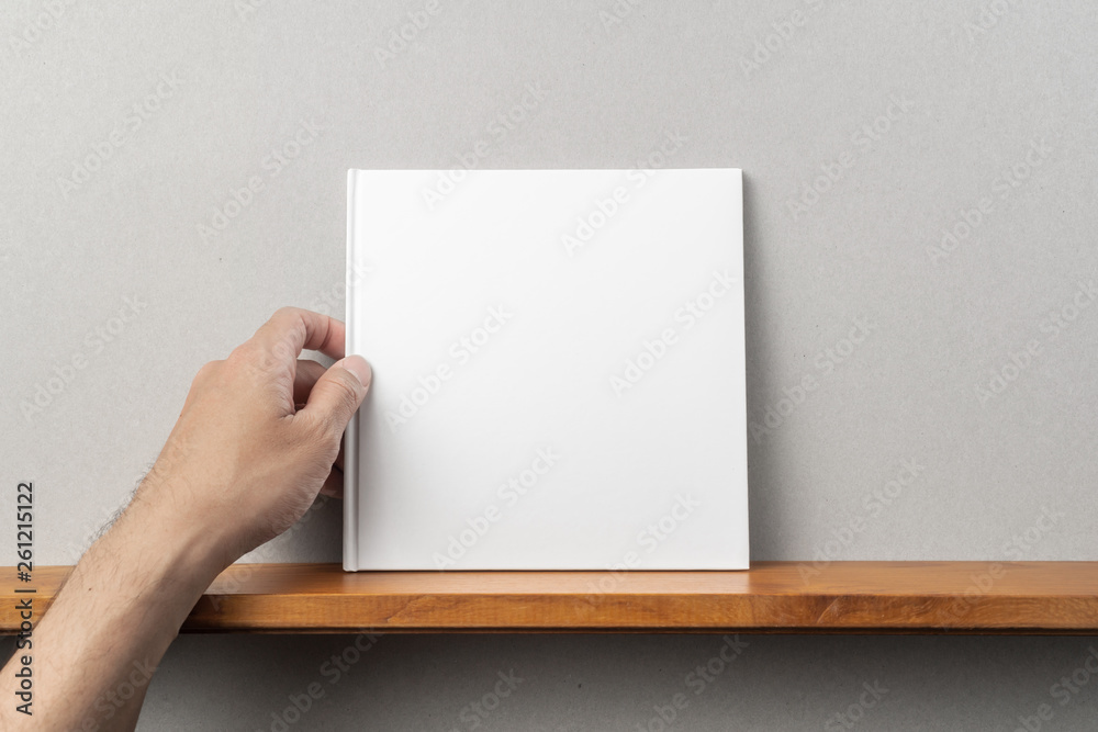 man's hand hold white square notebook on bookshelf