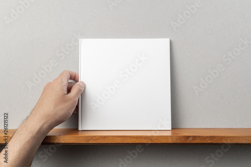 man's hand hold white square notebook on bookshelf
