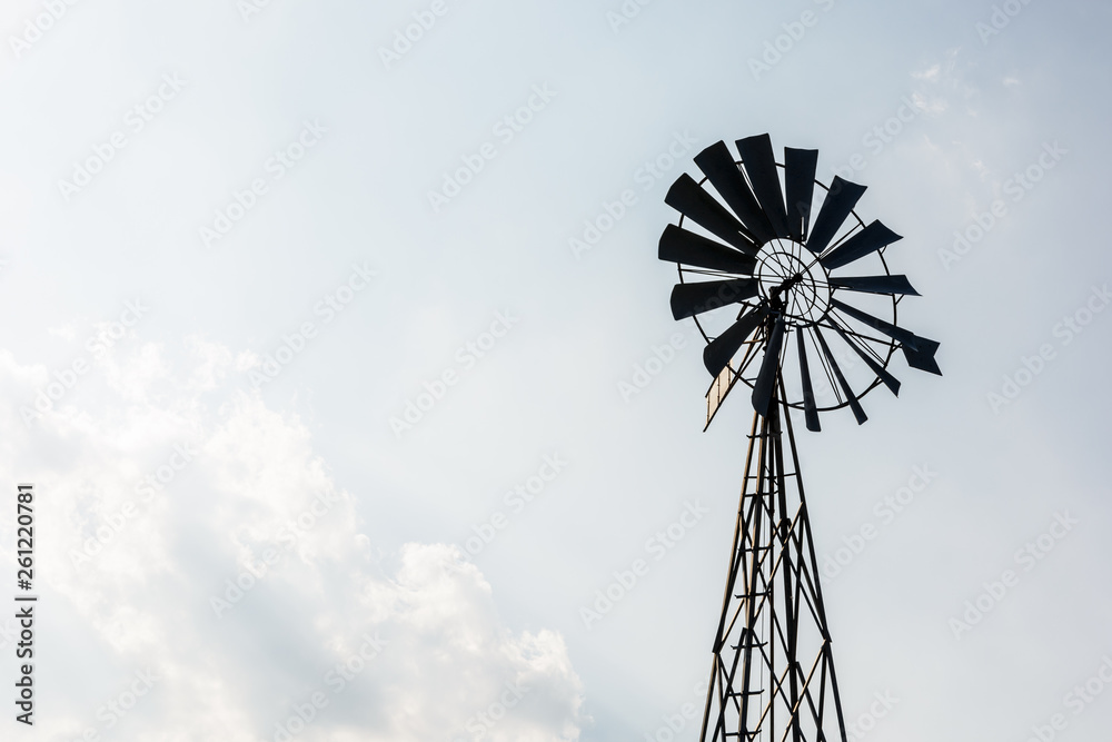 Low angle view of an old-fashioned, multi-bladed, metal wind powered water pump atop a lattice tower in backlight against a pale blue sky with white clouds. Low angle view of an old-fashioned, multi-bladed, metal wind powered water pump atop a lattice tower in backlight against a pale blue sky with white clouds.