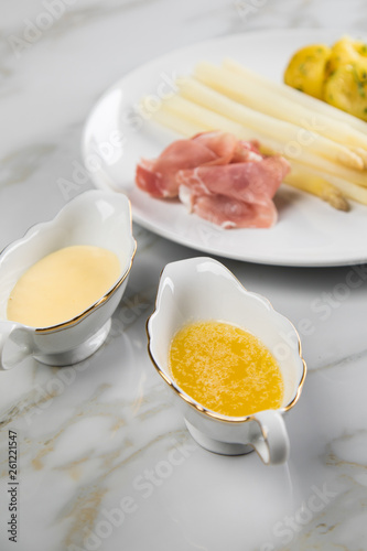 Weißer Spargel mit Sauce Hollandaise, zerlassener Butter, geräucherter Schinken und Kartoffeln auf Teller und Marmor Hintergrund