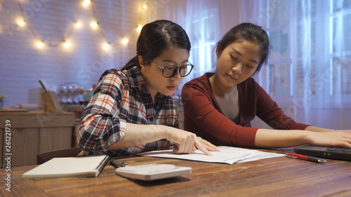 Focused young asian girl roommates checking analyzing utilities bills sitting together at kitchen wooden table. serious women reading bank loan documents financial planning expenses using laptop.