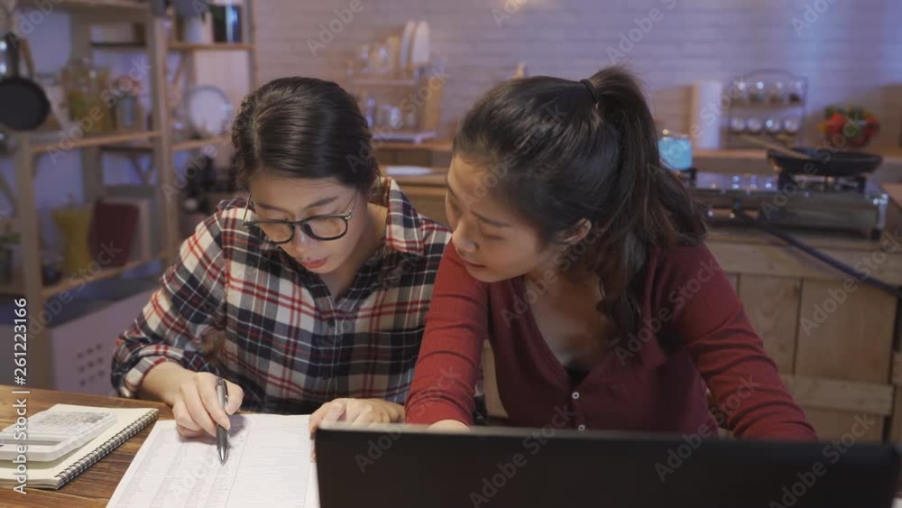young happy relaxed friends roommates at home kitchen table doing domestic debt accounting and calculating tax paperwork with bank paper and laptop computer. asian girls talking about month expense. young happy relaxed friends roommates at home kitchen table doing domestic debt accounting and calculating tax paperwork with bank paper and laptop computer. asian girls talking about month expense.