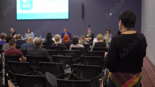 Business woman communicates with the speaker standing in the conference room among a group of people