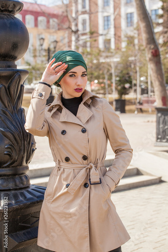 Stylish Muslim girl with a scarf on her head in a beige raincoat in the city. Stylish headband. The image of a modern woman in the city on the background of architecture