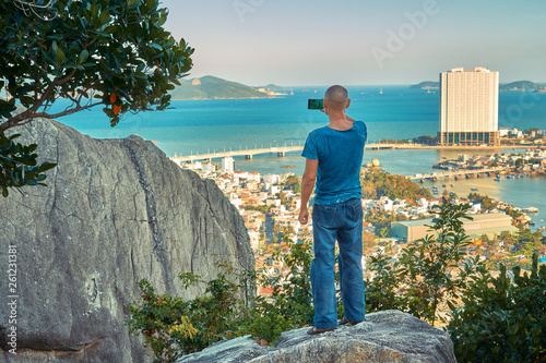Middle aged man hiker takes selfie from the rocky mountain viewpoint overlooking coastal city at sunset time