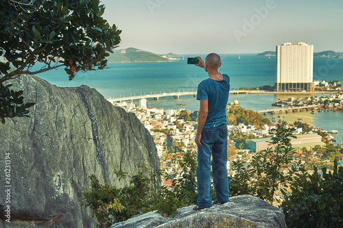 Middle aged man hiker takes selfie from the rocky mountain viewpoint overlooking coastal city at sunset time