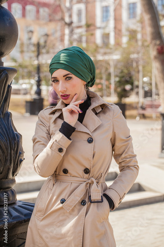 Stylish Muslim girl with a scarf on her head in a beige raincoat in the city. Stylish headband. The image of a modern woman in the city on the background of architecture