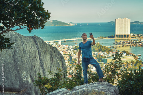 Middle aged man hiker takes selfie from the rocky mountain viewpoint overlooking coastal city at sunset time