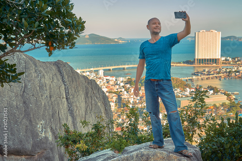 Middle aged man hiker takes selfie from the rocky mountain viewpoint overlooking coastal city at sunset time
