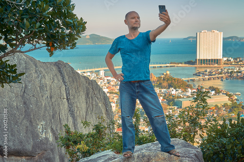 Middle aged man hiker takes selfie from the rocky mountain viewpoint overlooking coastal city at sunset time