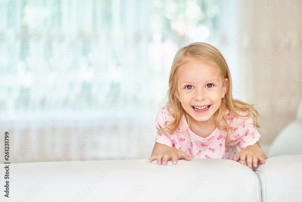Happy cute little Blonde girl, hide behind the white sofa in the room at home, portrait . Looking happily at the camera. Happy cute little Blonde girl, hide behind the white sofa in the room at home, portrait . Looking happily at the camera.