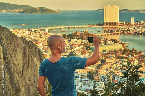 Middle aged man hiker takes selfie from the rocky mountain viewpoint overlooking coastal city at sunset time