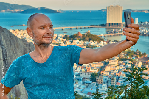 Middle aged man hiker takes selfie from the rocky mountain viewpoint overlooking coastal city at sunset time