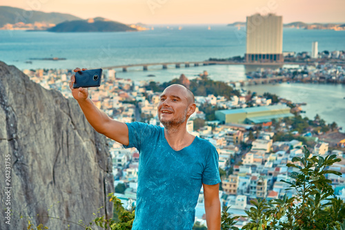 Middle aged man hiker takes selfie from the rocky mountain viewpoint overlooking coastal city at sunset time
