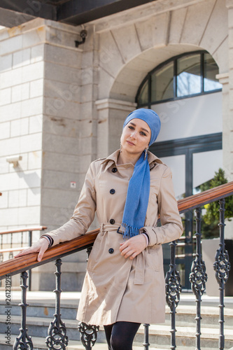 Stylish Muslim girl with a scarf on her head in a beige raincoat in the city. Stylish headband. The image of a modern woman in the city on the background of architecture