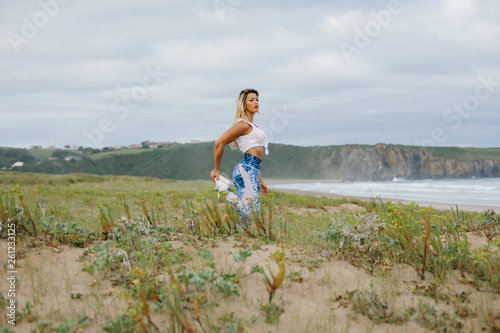 Fit muscular sporty young woman stretching quadriceps and exercising at the beach. Summer morning outdoor fitness workout. Playa de Xago, Asturias, Spain. photo