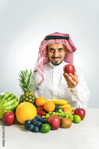 man from the Middle East in a traditional Arab national costume, stands in front of a mountain of fresh fruit, smiles joyfully and holds a red apple in his hand