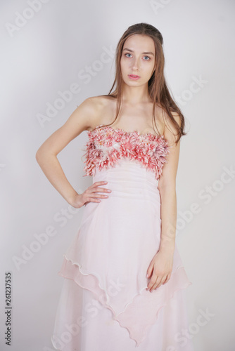 a charming young caucasian girl stands in a pink long prom dress with flower petals on her chest and poses on a white background in the Studio