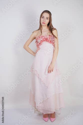 a charming young caucasian girl stands in a pink long prom dress with flower petals on her chest and poses on a white background in the Studio
