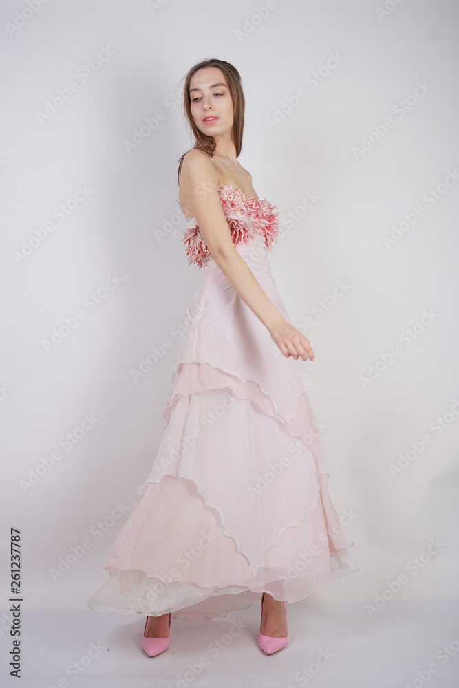 a charming young caucasian girl stands in a pink long prom dress with flower petals on her chest and poses on a white background in the Studio