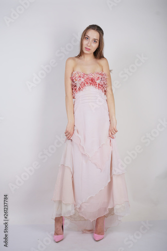 a charming young caucasian girl stands in a pink long prom dress with flower petals on her chest and poses on a white background in the Studio