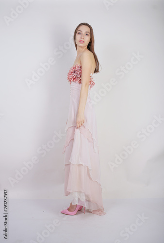 a charming young caucasian girl stands in a pink long prom dress with flower petals on her chest and poses on a white background in the Studio