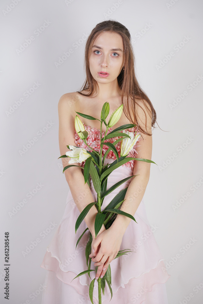 charming young caucasian girl in a pretty dress holds a lush Lily flower in her hands and stands on a white background in the Studio alone