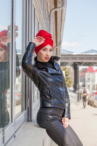 Stylish cheeky Muslim girl with a fashionably tied headscarf turban on her head in a red coat. Screaming image of a modern woman in the city against the backdrop of architecture